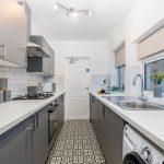 Modern galley kitchen with gray base cabinets, white countertops, left-side gas hob and oven, right-side sink by a window.