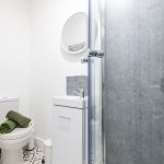 Compact bathroom with toilet, small white sink vanity, round mirror, and glass shower enclosure; green towels on the toilet and patterned floor tiles visible.