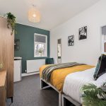 A bright modern bedroom with two single beds, mustard-yellow quilts, a dark green accent wall, a wooden wardrobe, and plants on a dresser left.