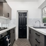 Narrow modern kitchen with dark gray cabinets, white countertops, gas stove on the left and a sink on the right, door at the far end, sunlight through a window on the right.