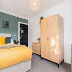 Bedroom with a mustard-yellow quilt, teal pillows, and a green accent wall above a white headboard.