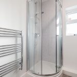 Curved glass corner shower with chrome-framed doors in a minimalist bathroom, marble-pattern walls visible behind the enclosure