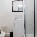 Compact modern bathroom featuring a curved glass shower enclosure, white vanity with sink, and a wall-mounted mirror above. Toilet partially visible on the left.