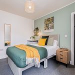 Cozy bedroom with green accent wall, bed with teal quilt, yellow throw, and mixed pillows; wooden nightstand, plants, and a black door nearby.