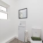 Bright, minimalist bathroom with a shower on the left, white vanity and cabinet, wall-mounted mirror, and a toilet with a green towel on top of the tank.