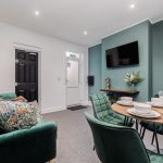 Cozy living room with emerald green chairs around a round dining table set with bowls and glasses, a wall-mounted TV on a teal wall, and decorative bust and vase of flowers nearby.